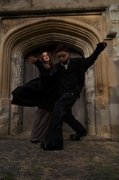 Two people dressed in dark, gothic-style clothing pose dramatically in front of an ornate archway made of stone. The setting appears historical, with intricate carvings above the wooden door and rough stone walls surrounding them. One person is wearing sunglasses and a chain necklace, striking a dynamic pose.