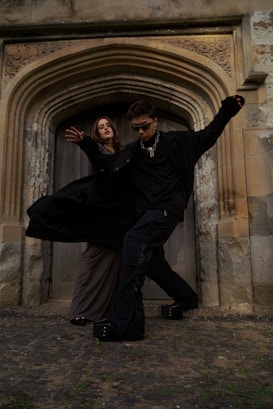 Two people dressed in dark, gothic-style clothing pose dramatically in front of an ornate archway made of stone. The setting appears historical, with intricate carvings above the wooden door and rough stone walls surrounding them. One person is wearing sunglasses and a chain necklace, striking a dynamic pose.