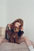A young person with long hair sits on a beige couch, playfully posing with a small, curly-haired brown dog. The person is wearing a cozy, oversized sweater with earthy tones and white socks. They hold the dog's ears up to resemble wings, adding a fun and whimsical element to the scene.