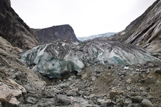 A large glacier with bluish-white ice sits between rocky cliffs. The glacier is surrounded by rubble and rocks, with the cliffs showing layers of dark stone. The sky is overcast, creating a stark contrast with the icy formations.