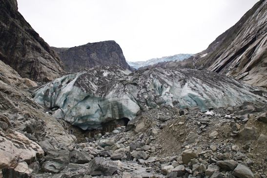 A large glacier with bluish-white ice sits between rocky cliffs. The glacier is surrounded by rubble and rocks, with the cliffs showing layers of dark stone. The sky is overcast, creating a stark contrast with the icy formations.