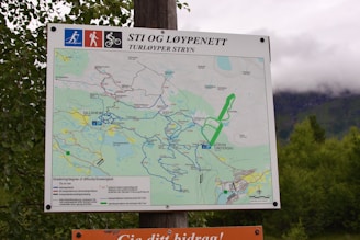 A map on a signboard detailing hiking and biking trails, located in an outdoor setting with lush greenery and mountains in the background. The map includes marked paths with various difficulty levels and parking areas, featuring labels in Norwegian. The sign is mounted on a wooden post.
