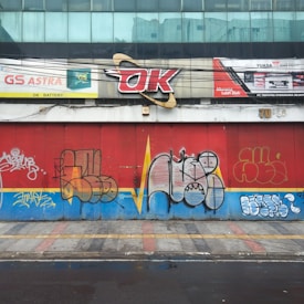 A city street scene featuring a building facade covered with colorful graffiti art. The wall is divided with red and blue paint, and multiple graffiti tags in varied styles and colors occupy the space. A sign above reads 'OK' with additional advertising for batteries, adding an urban commercial element. A tiled and slightly wet sidewalk is visible in the foreground.