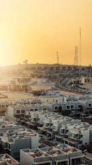 A suburban area with rows of modern, geometric white houses featuring rooftop amenities. In the background, a landscape of telecommunications towers and a satellite dish is visible against a bright yellow sky.
