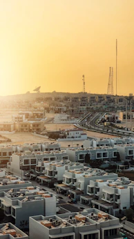 A suburban area with rows of modern, geometric white houses featuring rooftop amenities. In the background, a landscape of telecommunications towers and a satellite dish is visible against a bright yellow sky.