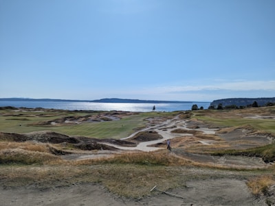 A coastal golf course with rolling sand dunes and patches of green grass, stretching towards a body of water. The backdrop features a vast sky and a distant shoreline.
