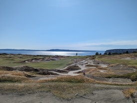 A coastal golf course with rolling sand dunes and patches of green grass, stretching towards a body of water. The backdrop features a vast sky and a distant shoreline.