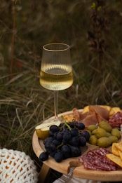 A glass of white wine stands on a grassy surface near a wooden platter filled with a variety of foods. The platter includes dark grapes, green olives, slices of cured meat, cheese, and half a pear. The setting appears to be outdoors, giving a rustic picnic vibe.