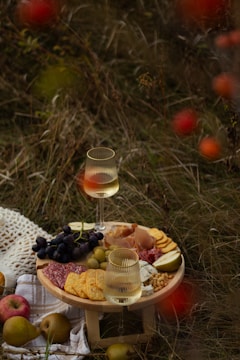 A rustic picnic setup on grass featuring a small wooden table with a charcuterie board. There are slices of salami, crackers, different cheeses, grapes, prosciutto, olives, and pear slices. Two glasses of white wine sit on the table. Apples and pears are scattered nearby, along with a crocheted cloth.