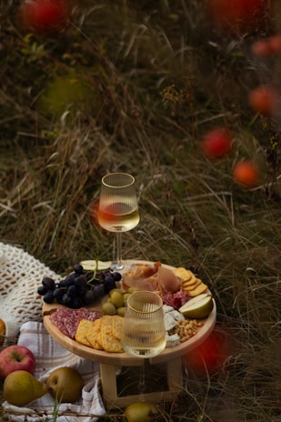 A rustic picnic setup on grass featuring a small wooden table with a charcuterie board. There are slices of salami, crackers, different cheeses, grapes, prosciutto, olives, and pear slices. Two glasses of white wine sit on the table. Apples and pears are scattered nearby, along with a crocheted cloth.