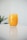 A glass filled with vibrant orange juice, placed on a reflective white surface. The background is blurred with a hint of green foliage, suggesting a plant.