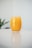 A glass filled with vibrant orange juice, placed on a reflective white surface. The background is blurred with a hint of green foliage, suggesting a plant.