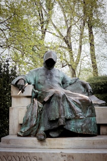 A bronze statue of a hooded figure sitting with a quill in hand, surrounded by lush greenery. The statue's face is obscured, adding an element of mystery.