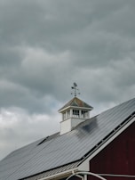 A rooftop with solar panels is visible beneath a cloudy sky. A weather vane sits atop a small cupola, adding a rustic element to the modern solar panels on the roof. The contrast between the dark solar panels and the white structure of the cupola is prominent.