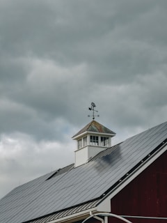 A rooftop with solar panels is visible beneath a cloudy sky. A weather vane sits atop a small cupola, adding a rustic element to the modern solar panels on the roof. The contrast between the dark solar panels and the white structure of the cupola is prominent.