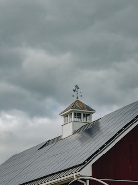 A rooftop with solar panels is visible beneath a cloudy sky. A weather vane sits atop a small cupola, adding a rustic element to the modern solar panels on the roof. The contrast between the dark solar panels and the white structure of the cupola is prominent.