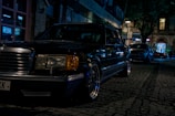 A classic luxury car is parked on a dimly lit cobblestone street at night. The car is a black sedan, with chrome detailing and distinctive rectangular headlights. Surrounding it are softly lit storefronts and other parked cars, creating an urban evening atmosphere.