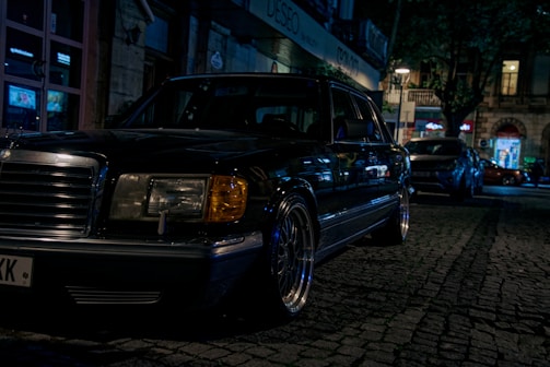 A classic luxury car is parked on a dimly lit cobblestone street at night. The car is a black sedan, with chrome detailing and distinctive rectangular headlights. Surrounding it are softly lit storefronts and other parked cars, creating an urban evening atmosphere.