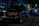 A classic luxury car is parked on a dimly lit cobblestone street at night. The car is a black sedan, with chrome detailing and distinctive rectangular headlights. Surrounding it are softly lit storefronts and other parked cars, creating an urban evening atmosphere.