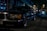 A classic luxury car is parked on a dimly lit cobblestone street at night. The car is a black sedan, with chrome detailing and distinctive rectangular headlights. Surrounding it are softly lit storefronts and other parked cars, creating an urban evening atmosphere.