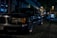 A classic luxury car is parked on a dimly lit cobblestone street at night. The car is a black sedan, with chrome detailing and distinctive rectangular headlights. Surrounding it are softly lit storefronts and other parked cars, creating an urban evening atmosphere.
