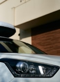 A close-up of a car's headlight, showing the sleek design of the light embedded in the vehicle's front. The background includes part of a building with cream and brown walls.