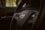 A close-up of a car steering wheel with a Hyundai logo in the center. The image highlights the steering wheel's texture and intricate details, with visible buttons for controls.