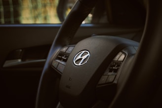 A close-up of a car steering wheel with a Hyundai logo in the center. The image highlights the steering wheel's texture and intricate details, with visible buttons for controls.