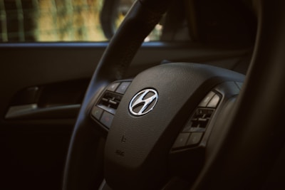 A close-up of a car steering wheel with a Hyundai logo in the center. The image highlights the steering wheel's texture and intricate details, with visible buttons for controls.