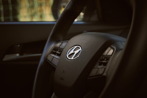 A close-up of a car steering wheel with a Hyundai logo in the center. The image highlights the steering wheel's texture and intricate details, with visible buttons for controls.