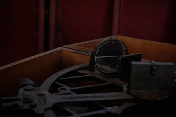 A vintage navigational instrument is positioned inside a worn wooden box. The instrument, possibly an octant or sextant, features arcs and a circular mirror, suggesting its use for celestial navigation. The dim lighting and shadowy ambiance evoke a sense of history and maritime exploration.