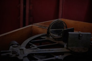 A vintage navigational instrument is positioned inside a worn wooden box. The instrument, possibly an octant or sextant, features arcs and a circular mirror, suggesting its use for celestial navigation. The dim lighting and shadowy ambiance evoke a sense of history and maritime exploration.
