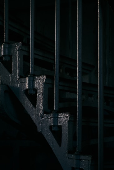 Dark, industrial-style staircase with metal railings and steps. The lighting creates a moody atmosphere, emphasizing shadows and the textured surface of the metal.