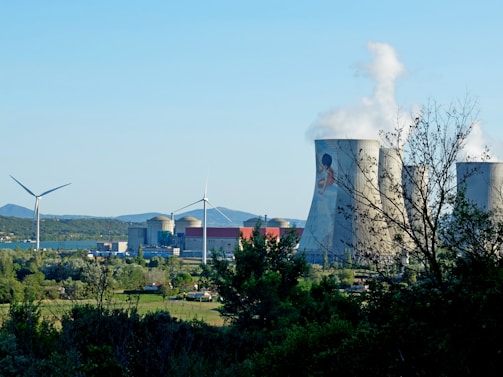 A landscape featuring a power plant with large cooling towers emitting steam and two wind turbines. The foreground is filled with trees and greenery, while the background displays a clear blue sky and distant hills.