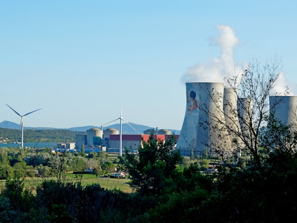 A landscape featuring a power plant with large cooling towers emitting steam and two wind turbines. The foreground is filled with trees and greenery, while the background displays a clear blue sky and distant hills.