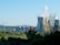 A landscape featuring a power plant with large cooling towers emitting steam and two wind turbines. The foreground is filled with trees and greenery, while the background displays a clear blue sky and distant hills.