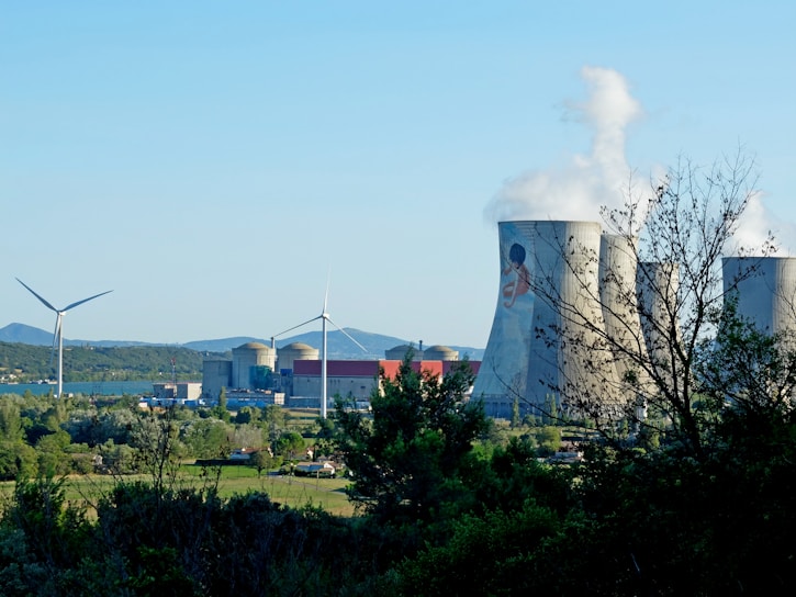 A landscape featuring a power plant with large cooling towers emitting steam and two wind turbines. The foreground is filled with trees and greenery, while the background displays a clear blue sky and distant hills.