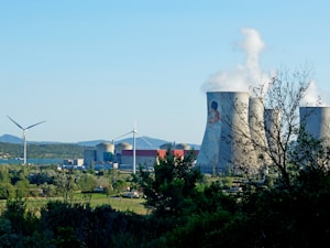 A landscape featuring a power plant with large cooling towers emitting steam and two wind turbines. The foreground is filled with trees and greenery, while the background displays a clear blue sky and distant hills.