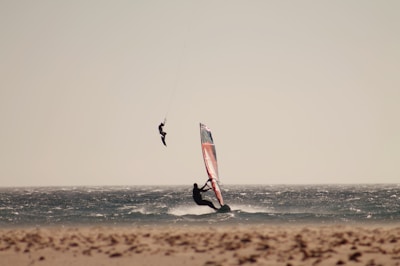 A person is windsurfing on the ocean, while another person is kiteboarding nearby, suspended in mid-air. The scene captures the dynamic motion and excitement of both water sports against a clear, expansive sky.