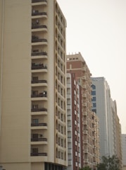 A row of high-rise residential buildings with multiple balconies and windows. The architecture features geometrical patterns, with one building having arches on the facade. The structures have varying colors, including beige, red, and white. The sky appears clear and the trees are visible at the bottom of the image.