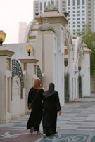 Two individuals dressed in black abayas walk down a paved area bordered by architectural elements featuring arches and columns. Above them, ornate lanterns provide lighting. In the background, modern skyscrapers rise into the hazy sky, partially obscured by trees on the right.