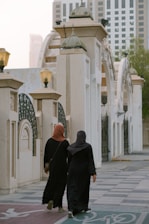 Two individuals dressed in black abayas walk down a paved area bordered by architectural elements featuring arches and columns. Above them, ornate lanterns provide lighting. In the background, modern skyscrapers rise into the hazy sky, partially obscured by trees on the right.