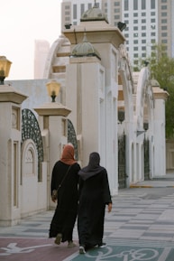 Two individuals dressed in black abayas walk down a paved area bordered by architectural elements featuring arches and columns. Above them, ornate lanterns provide lighting. In the background, modern skyscrapers rise into the hazy sky, partially obscured by trees on the right.