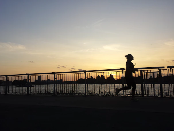 Sunset silhouette of a lone runner sprinting along a riverside path, blue sky fading.