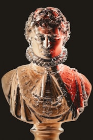 A glass sculpture of a historical figure, adorned in detailed attire including a prominent ruff collar. The sculpture features intricate facial details with a glossy finish and is illuminated with warm tones.