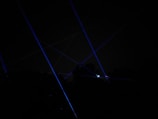 Blue laser beams crisscross against a dark night sky, creating a dramatic and futuristic effect. The beams appear to originate from multiple points, possibly placed on the ground or on structures, and some foliage is faintly visible at the bottom of the image.