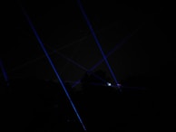 Blue laser beams crisscross against a dark night sky, creating a dramatic and futuristic effect. The beams appear to originate from multiple points, possibly placed on the ground or on structures, and some foliage is faintly visible at the bottom of the image.