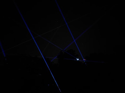 Blue laser beams crisscross against a dark night sky, creating a dramatic and futuristic effect. The beams appear to originate from multiple points, possibly placed on the ground or on structures, and some foliage is faintly visible at the bottom of the image.