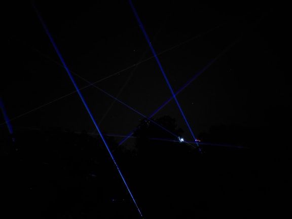 Blue laser beams crisscross against a dark night sky, creating a dramatic and futuristic effect. The beams appear to originate from multiple points, possibly placed on the ground or on structures, and some foliage is faintly visible at the bottom of the image.