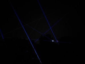 Blue laser beams crisscross against a dark night sky, creating a dramatic and futuristic effect. The beams appear to originate from multiple points, possibly placed on the ground or on structures, and some foliage is faintly visible at the bottom of the image.